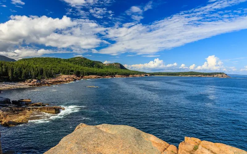 Large puffy clouds dot a brilliant blue sky as wave crash against the rocky coastline of Acadia.