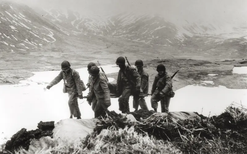 Six men with guns carry a person on a stretcher in a valley.