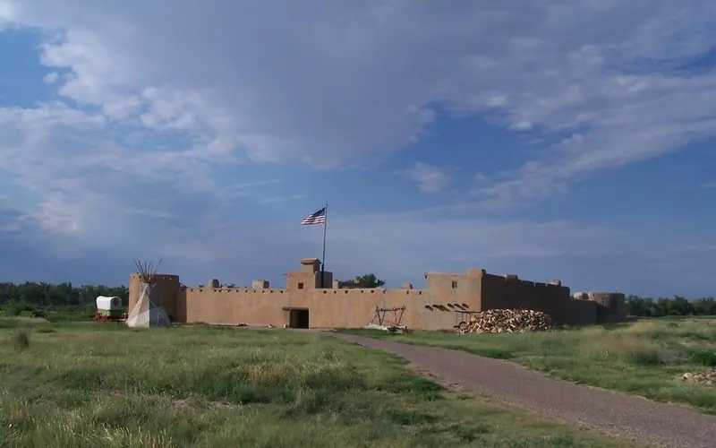 Bent's Old Fort with shortgrass prairie, tepee and wagon in foreground and blue sky & clouds above