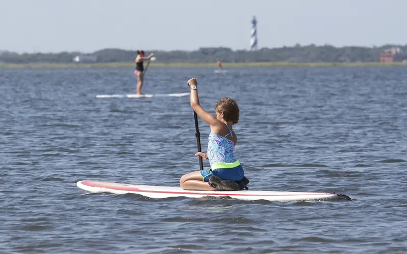 A young girl paddles across the water with the Cape Hatteras Lighthouse visible in the distance.