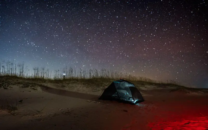 A red light illuminates a tent on the sand, with a star filled sky behind it.