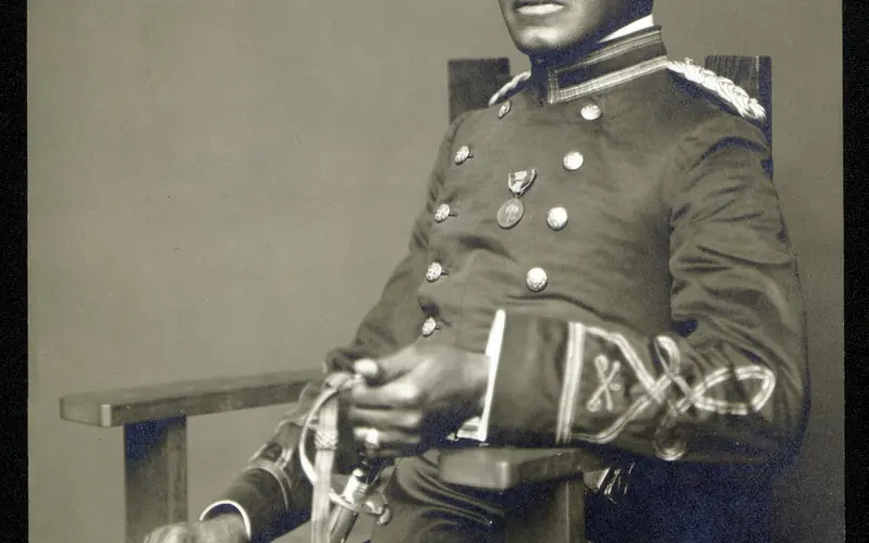 An African American man in a military uniform holding a sabre and wearing a cap sitting in a chair