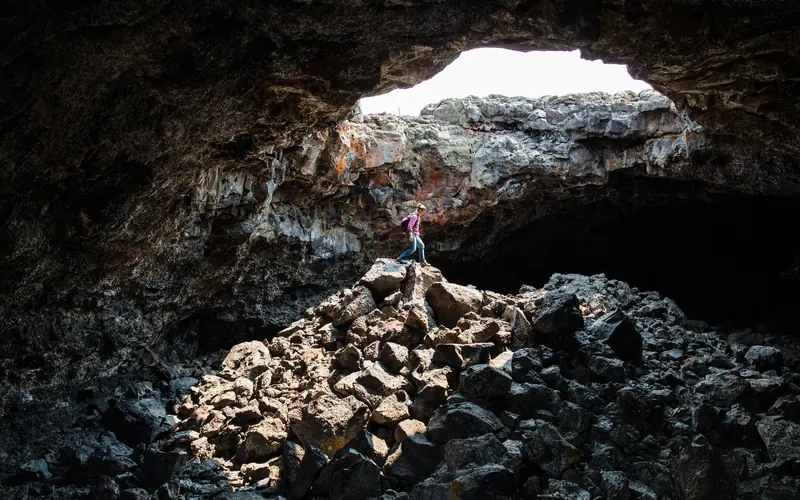 A figure stands inside a large lava tube on a pile of rubble, lit by a round opening overhead.
