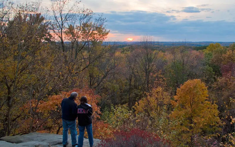 Two people stand at the edge of a gray rock cliff; in the distance, sunset and trees in fall colors.