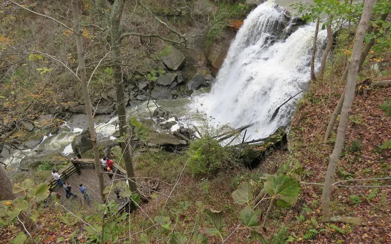 Viewed from above, people stand on a platform near frothy white water falling over a rocky ledge.