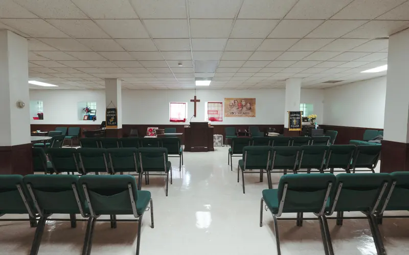 Rows of green seats in a large white room pointed toward a pulpit and a cross on a wall.