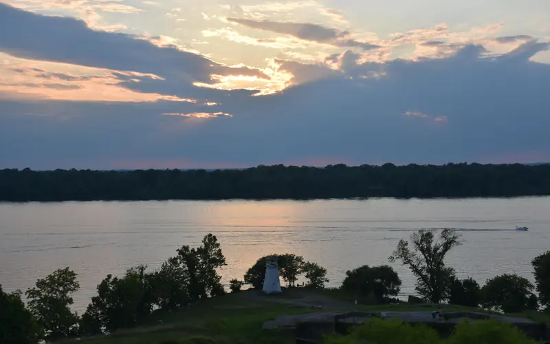 a green shoreline with small lighthouse along river at sunset