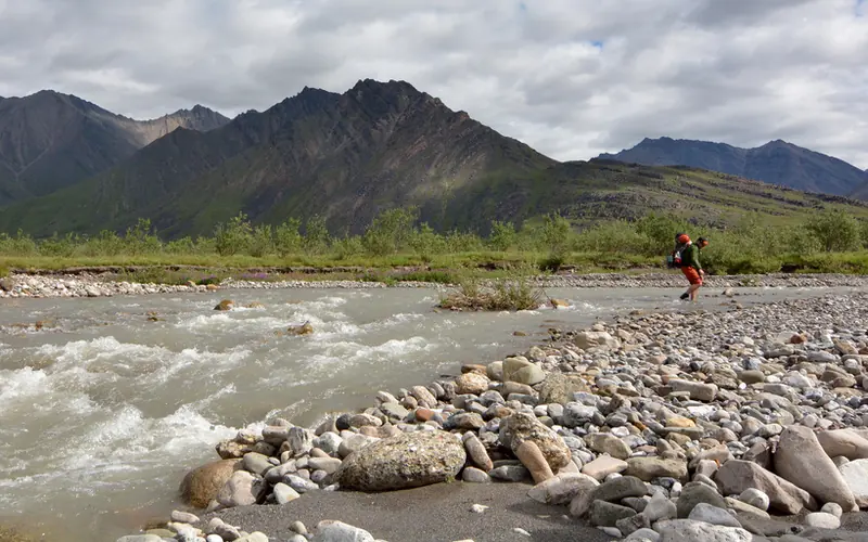 A hiker crosses a stream with mountains in the background