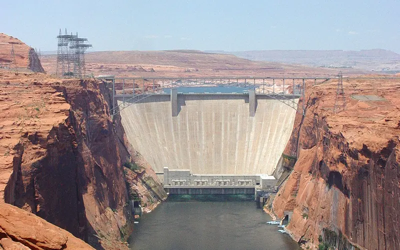 A concrete dam plugs high canyon walls. Buildings and wires on the sides.