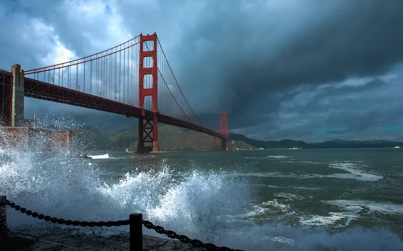 Orange Golden Gate Bridge with waves crashing in foreground and storm clouds behind.
