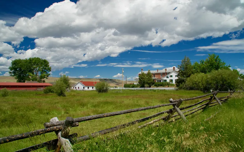 Fenced green pasture in front, 3 ranch buildings across center, blue sky with large white clouds.