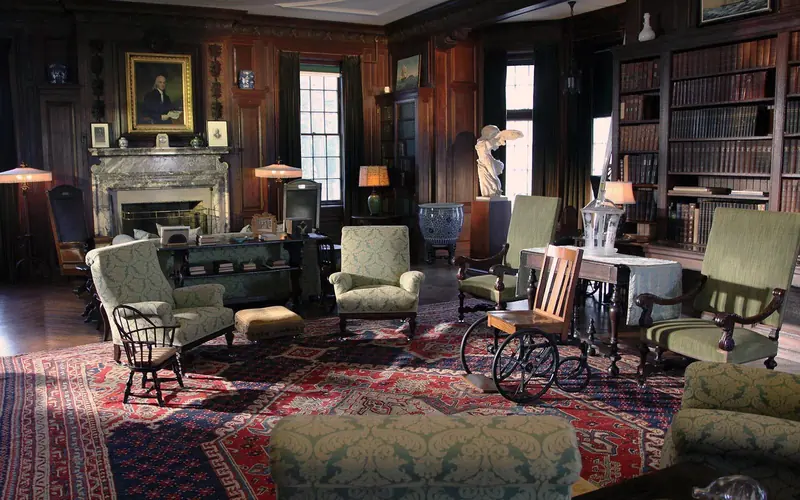 A richly furnished room with bookcases and fireplace with portrait hanging above.