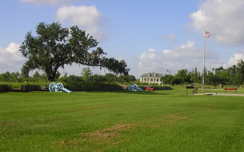 An oak tree arches over a cannon-guarded rampart with historic house and American flag in background
