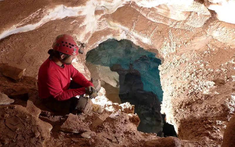 A cave explorer sits on some rocks and looks at a clear blue lake in Jewel Cave.