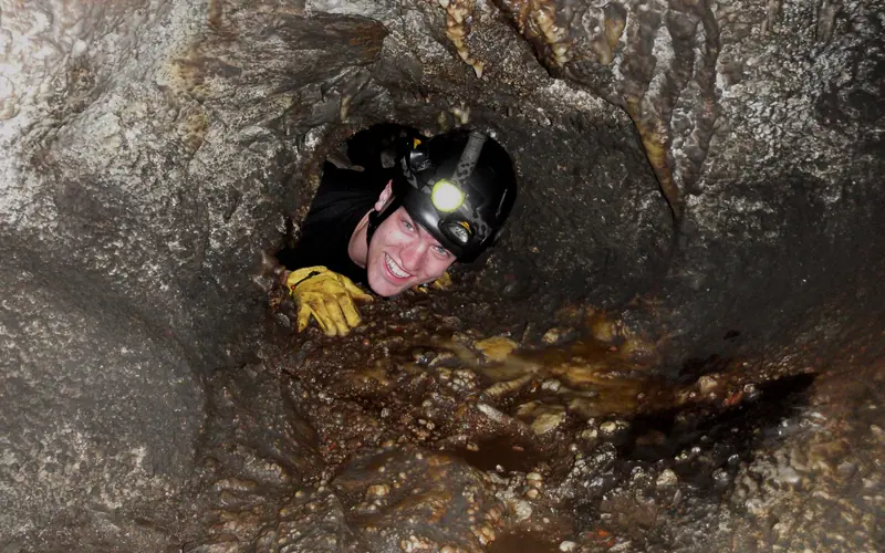 A caver squeezes through a very small and tight opening, surrounded by multi-colored rocks.