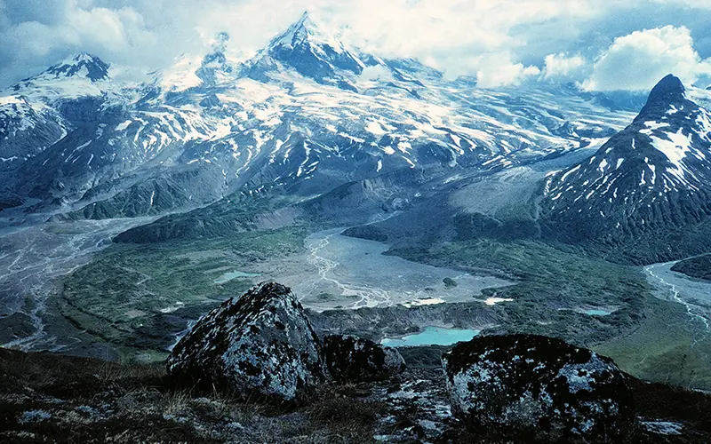 Photo of a stratovolcano flanked with glaciers towering over a river valley.