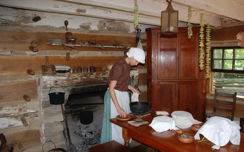 Park ranger is period dress standing near fireplace, cooking in the cabin.