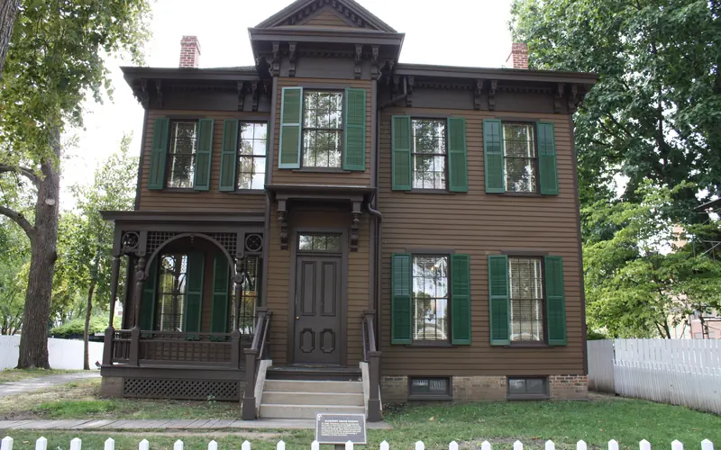 2 story brown house with dark green shutters and white fence.