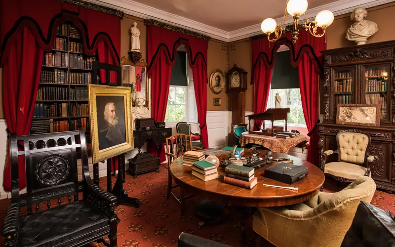 View of a study with a round center table cluttered with books and writing implements.