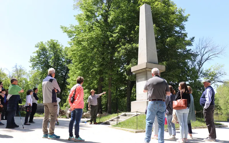 A park ranger stands in front of a stone obelisk monument pointing and talking to visitors