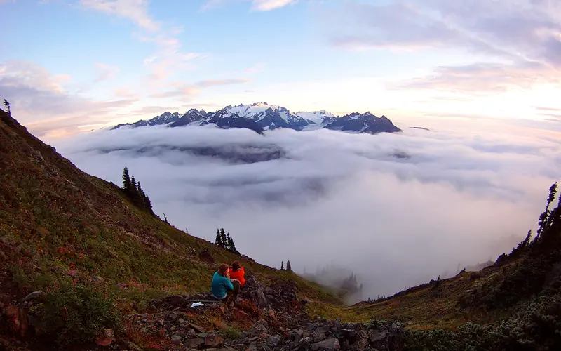 Hikers sit and watch the sun set behind snow-capped mountains.