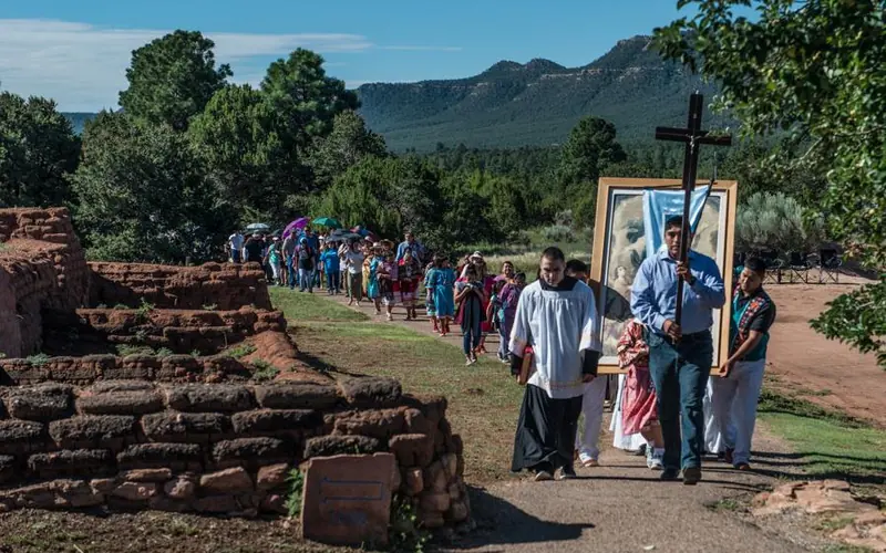 A line of people walk past adobe bricks, trees, and the road.
