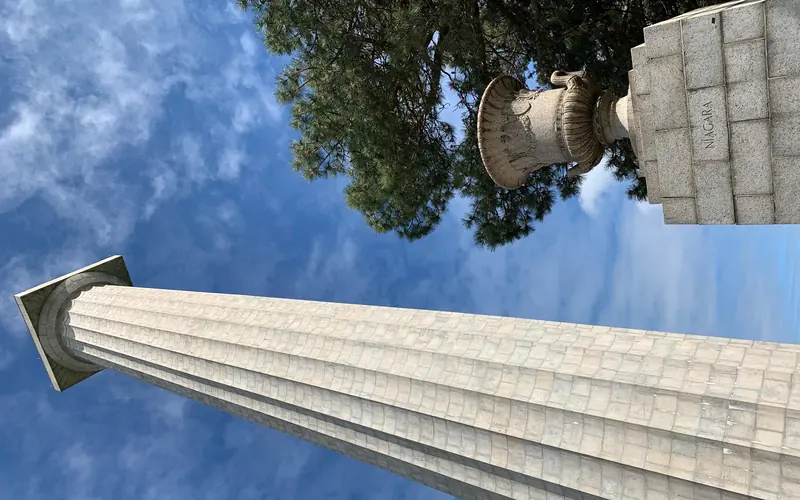 352 foot tall stone memorial column rises up to touch blue sky. In front a 5' urn on pedestal