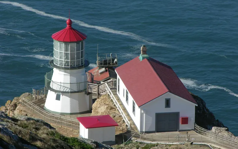 A three-story-tall, white-sided, red-roofed lighthouse adjacent to three other small buildings.