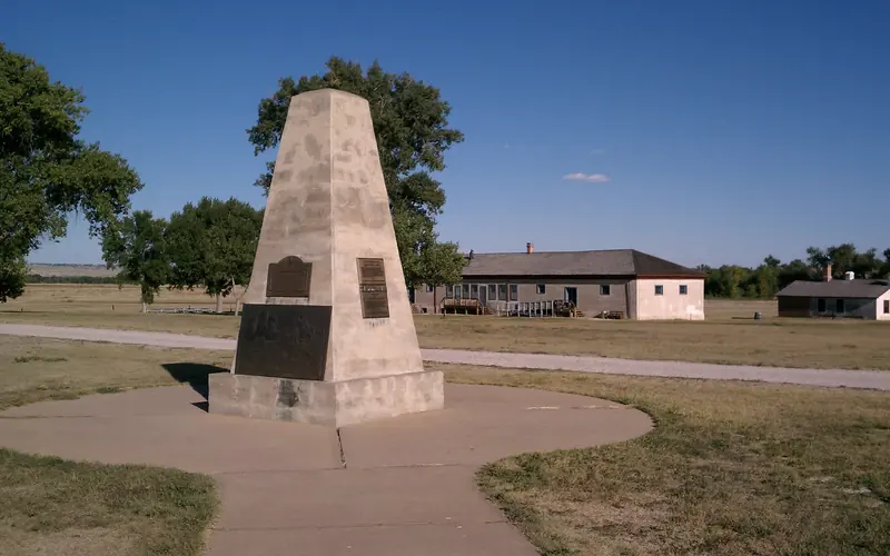 A tall obelisk monument with bronze plaques sits on a paved pathway in front of a building.