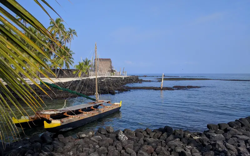 A double-hulled canoe sits in Keoneʻele Cove with Hale o Keawe in the background.