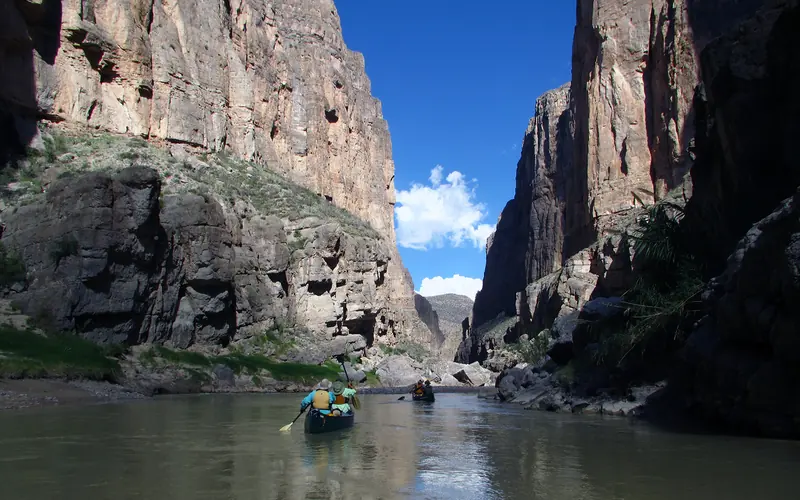 People in two canoes paddle on a river threading through high canyon walls.