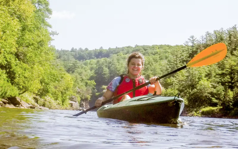 A woman paddles a kayak past cliffs on a forested river.