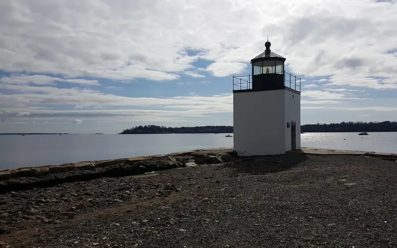 A white rectangular lighthouse approximately 20 feet tall is near the water on a gravel road.