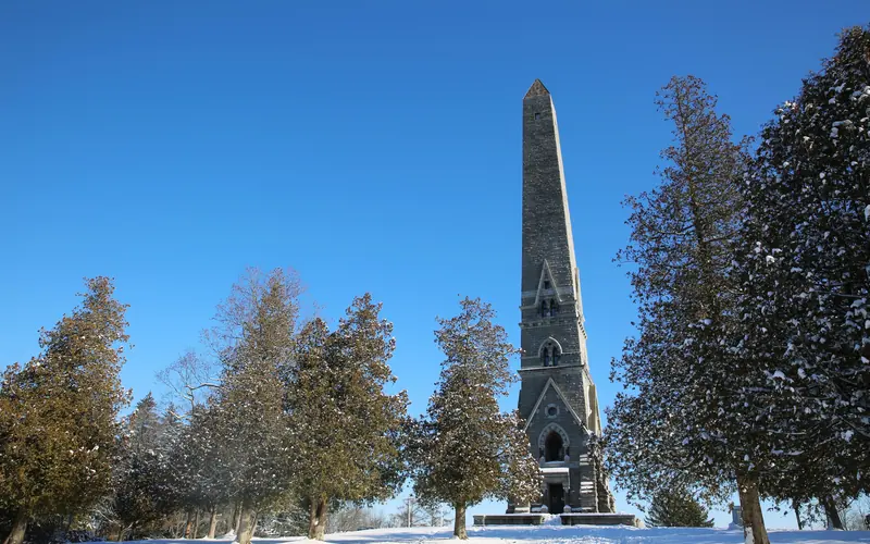 A narrow, stone obelisk on top of a snowy hill reaches into a clear sky.