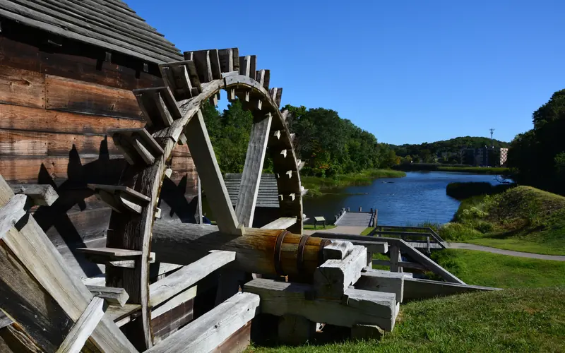 Approximately 15' by 15' spoked wooden wheel beside river and under blue sky.
