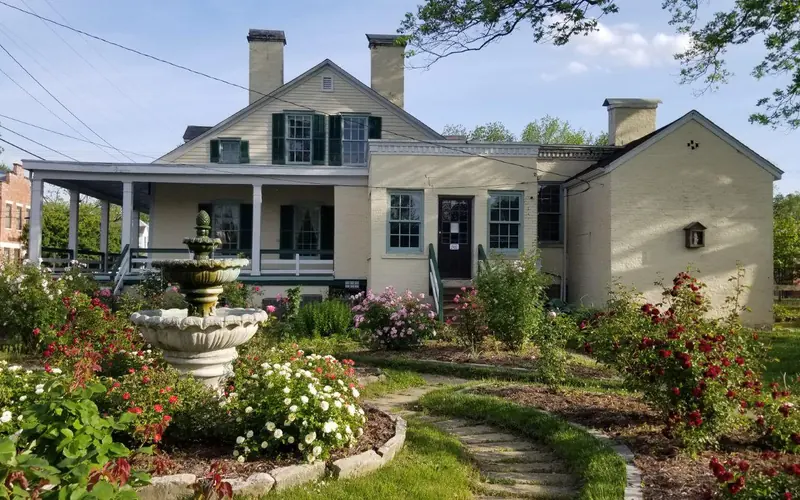 Flower beds with blooming flowers and a fountain, with a cream-colored house behind.