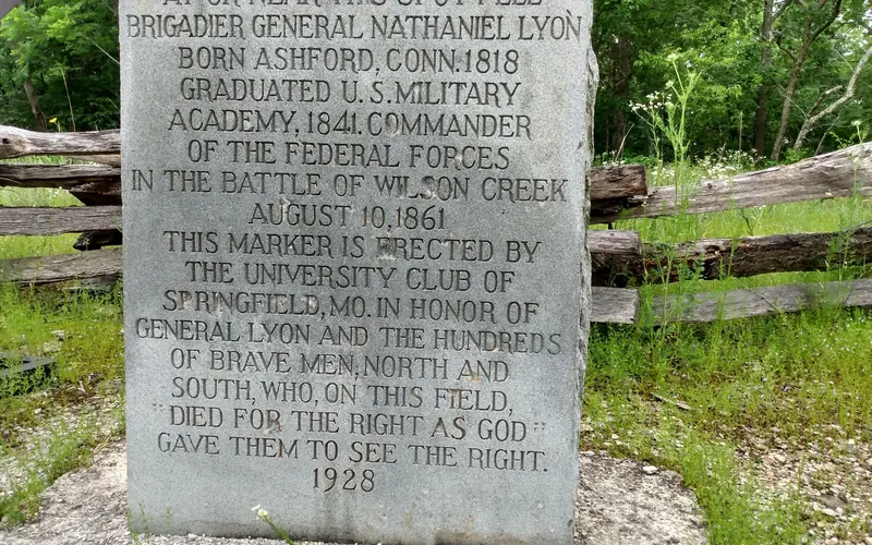 A stone marker with engraving sits in front of a wooden fence and trees