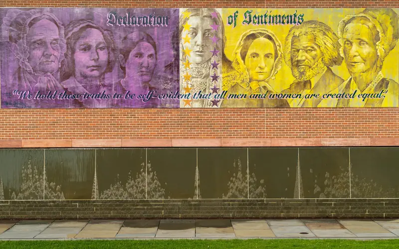 A gold and purple mural featuring faces of historic figures, over a stone wall.