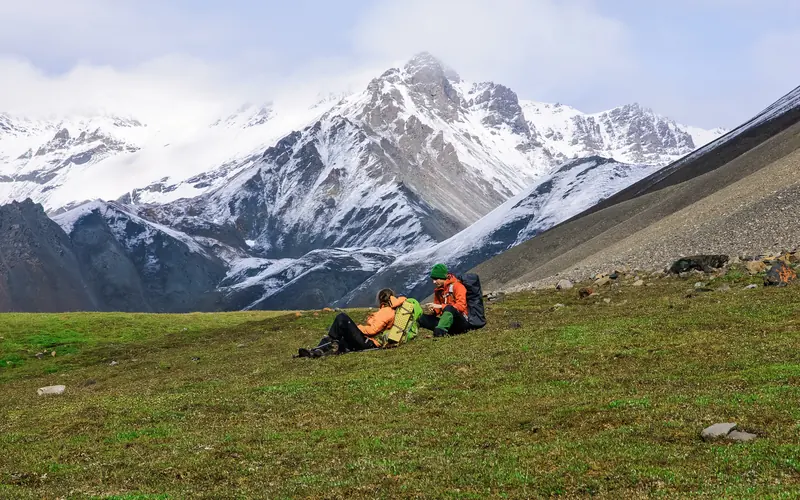 Two backpackers sitting in an alpine meadow with snowy mountains in the background