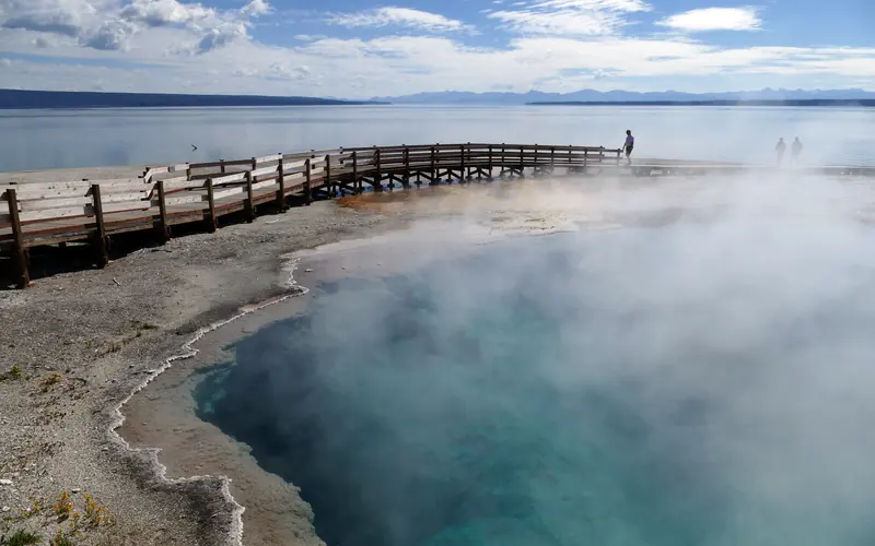 A visitor stands on a boardwalk near a hot spring and a lake.