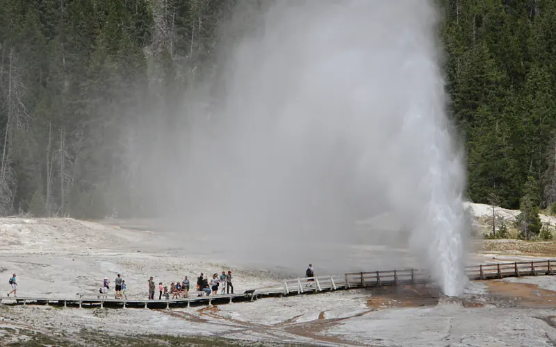 People on a wooden boardwalk watch a geyser erupt.